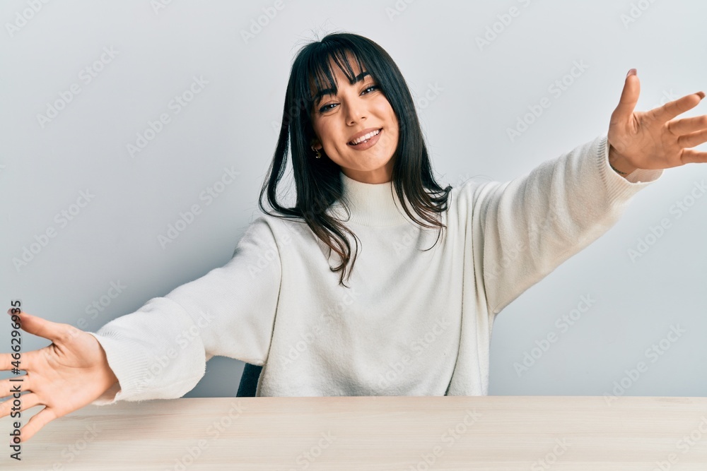 Young brunette woman with bangs wearing casual clothes sitting on the table looking at the camera smiling with open arms for hug. cheerful expression embracing happiness. Young brunette woman with bangs wearing casual clothes sitting on the table looking at the camera smiling with open arms for hug. cheerful expression embracing happiness.