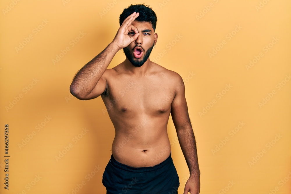 Arab man with beard wearing swimwear shirtless doing ok gesture shocked with surprised face, eye looking through fingers. unbelieving expression.