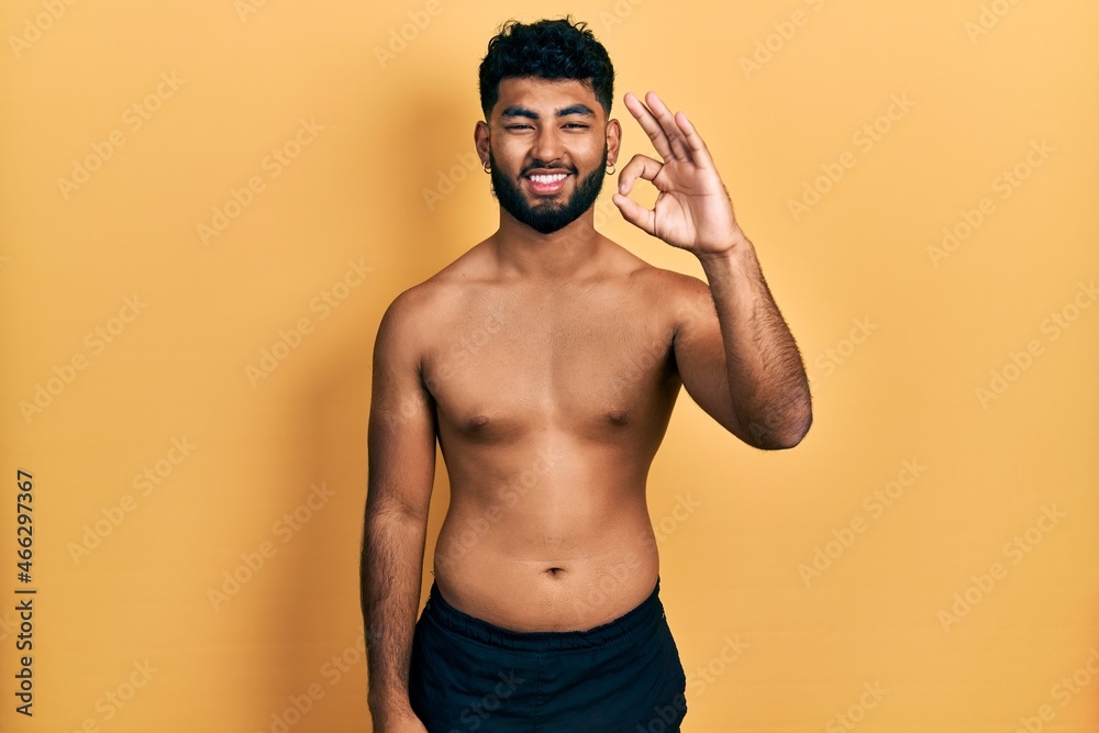 Arab man with beard wearing swimwear shirtless smiling positive doing ok sign with hand and fingers. successful expression.