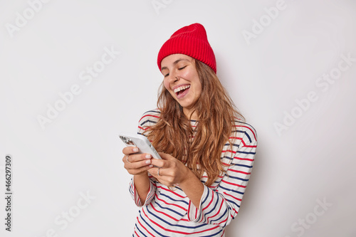 Cheerful millennial girl uses modern mobile phone sends text messages chats online wears red hat striped jumper has piercing in nose isolated over white background. People and technology concept