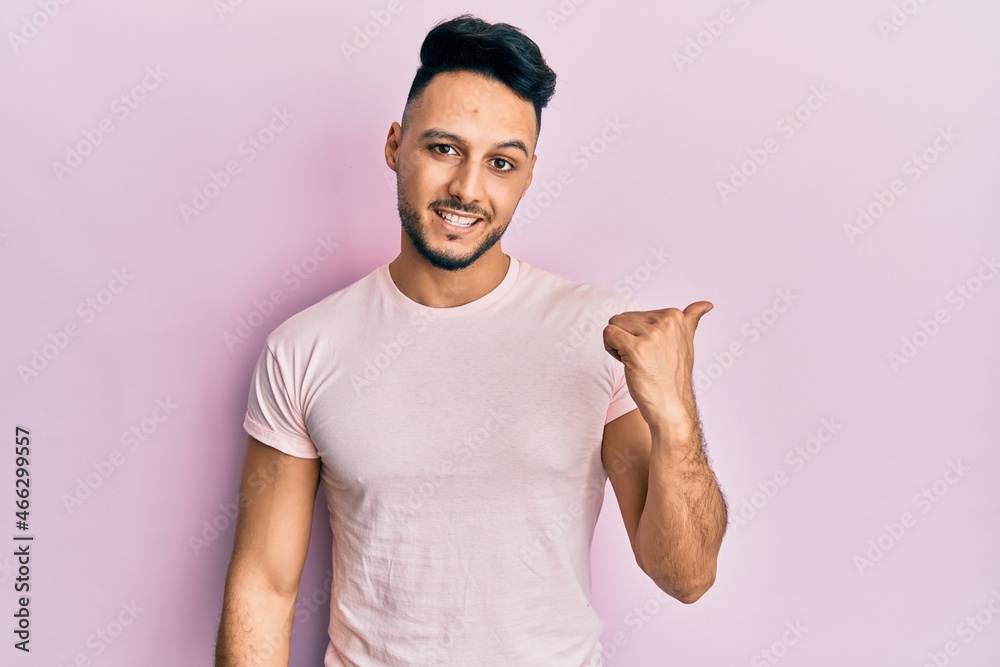 Young arab man wearing casual clothes smiling with happy face looking and pointing to the side with thumb up. Young arab man wearing casual clothes smiling with happy face looking and pointing to the side with thumb up.