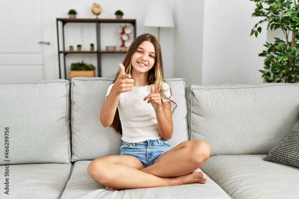 Young brunette teenager sitting on the sofa at home pointing fingers to camera with happy and funny face. good energy and vibes. Young brunette teenager sitting on the sofa at home pointing fingers to camera with happy and funny face. good energy and vibes.