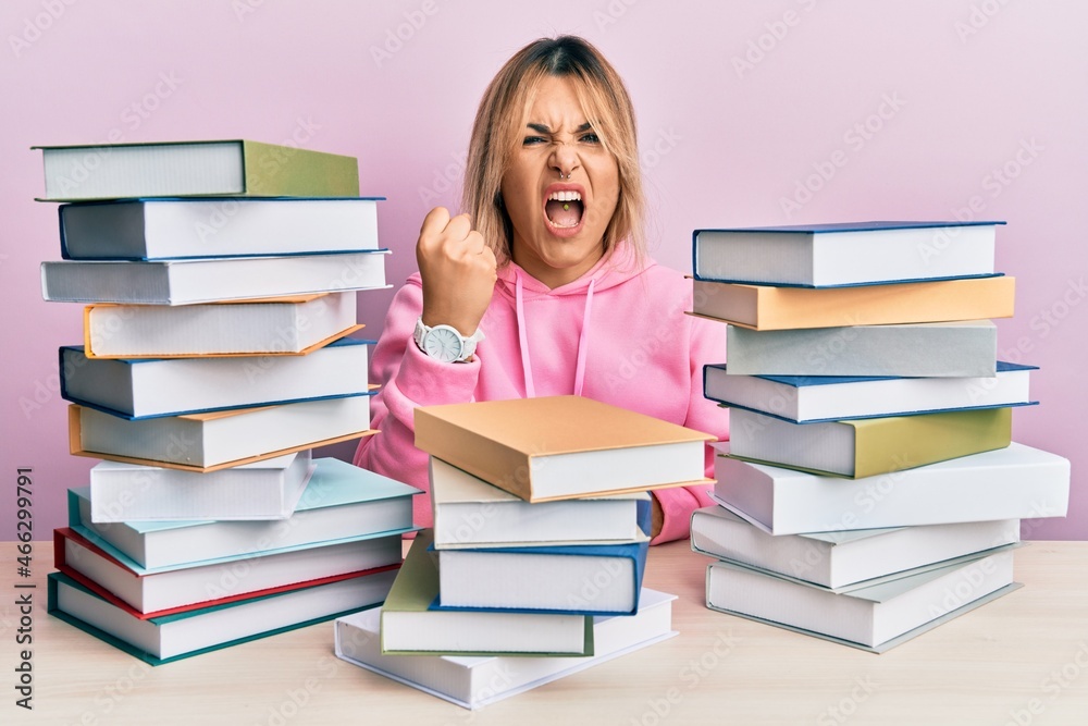 Young caucasian woman sitting on the table with books angry and mad raising fist frustrated and furious while shouting with anger. rage and aggressive concept. Young caucasian woman sitting on the table with books angry and mad raising fist frustrated and furious while shouting with anger. rage and aggressive concept.