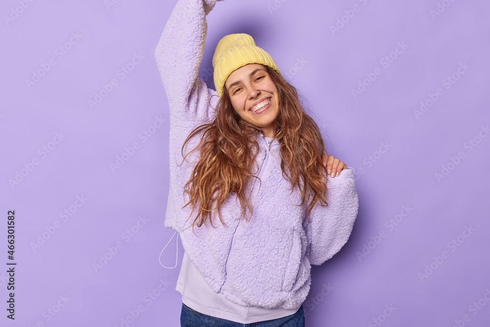 Happy young pretty woman raises arm up tilts head and smiles gladfully dressed in stylish outerwear has piercing in nose celebrates triumph isolated over purple background feels free and joyful Happy young pretty woman raises arm up tilts head and smiles gladfully dressed in stylish outerwear has piercing in nose celebrates triumph isolated over purple background feels free and joyful