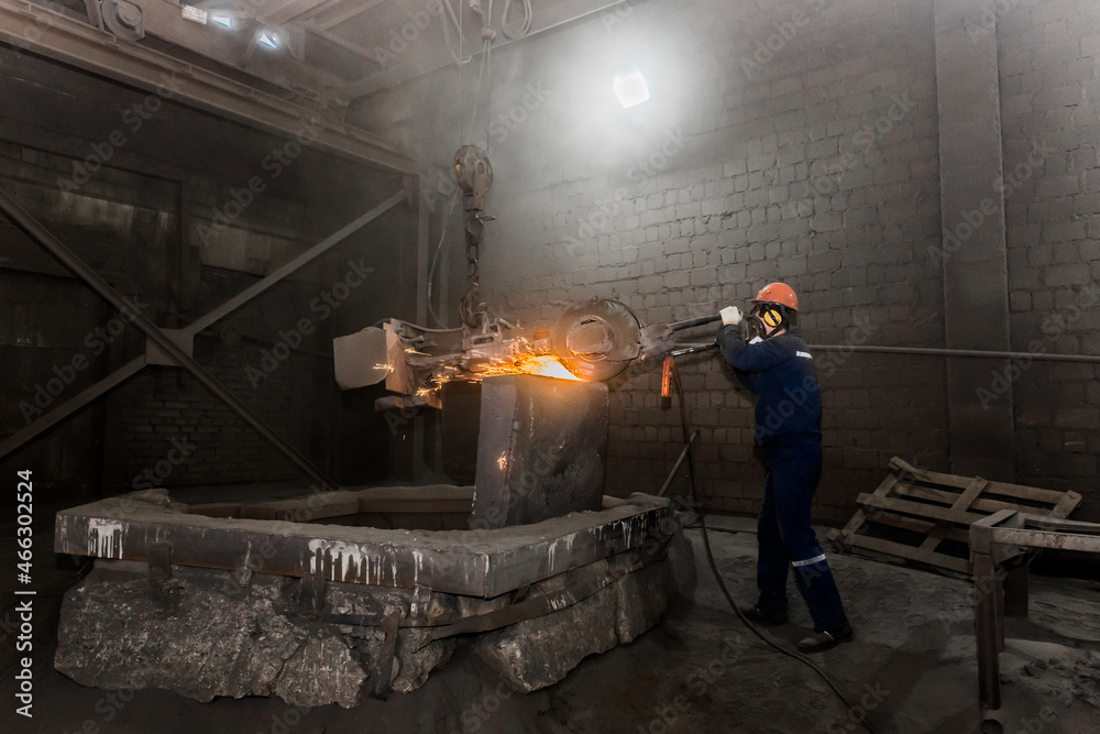 A male worker in a protective helmet, respirator, overalls manages heavy grinding equipment for cast iron concrete tubing with flying sparks in the workshop of an industrial plant