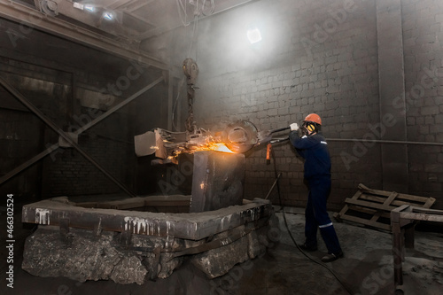 A male worker in a protective helmet, respirator, overalls manages heavy grinding equipment for cast iron concrete tubing with flying sparks in the workshop of an industrial plant