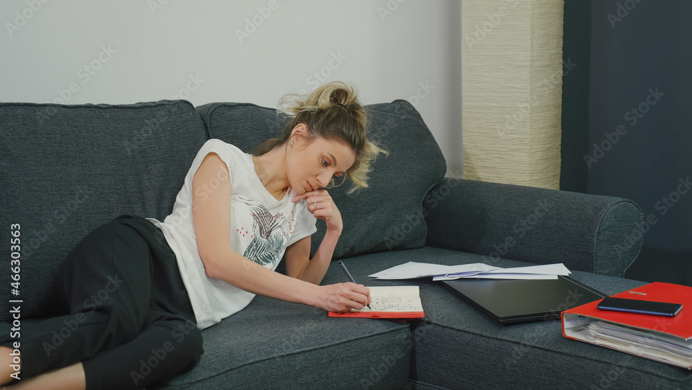 Woman makes notes in notebook while lying on couch at home in a modern apartment. Woman makes notes in notebook while lying on couch at home in a modern apartment.