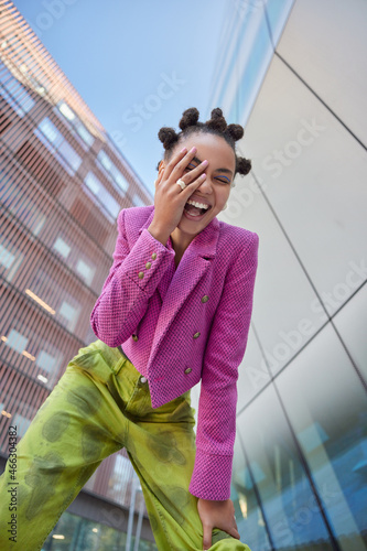 View from below of overjoyed female model keeps hand on face feels very happy wears stylish pink jacket and green trouses spends free time in urban place expresses positive emotions. Youth style photo