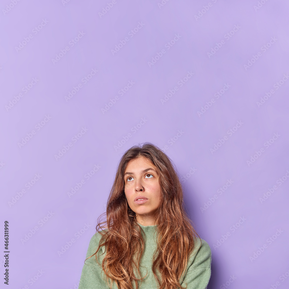 Naklejka premium Vertical image of thoughtful long haired woman has bored unhappy expression focused above thinks about something wears casual jumper isolated over purple background with blank space for your text