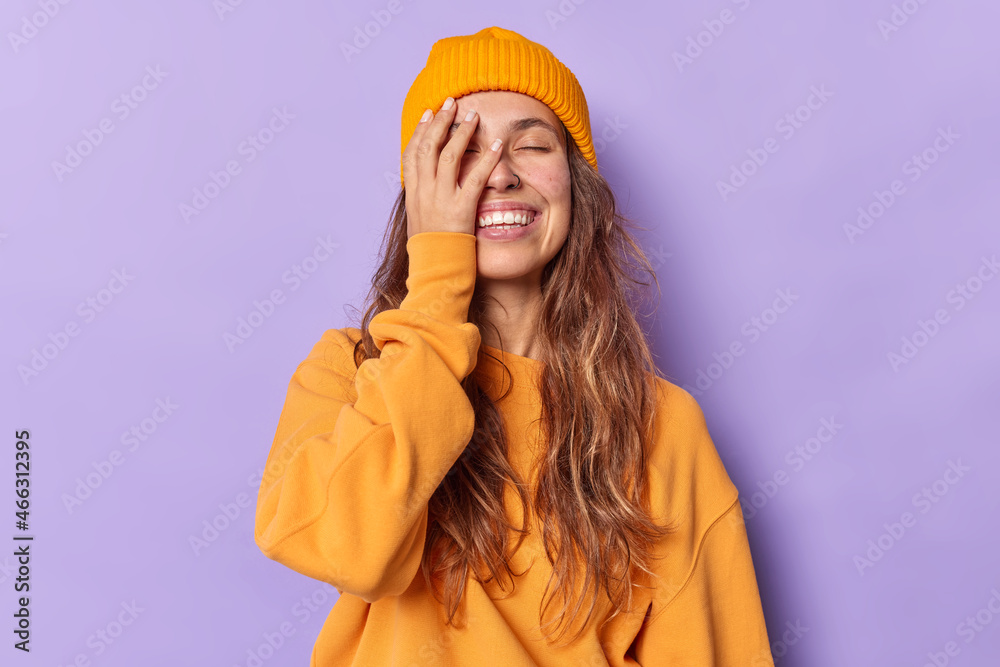Positive sincere millennial girl has long hair makes face palm keeps eyes closed has upbeat mood wears casual orange jumper and hat isolated over purple background. People emotions feelings concept Positive sincere millennial girl has long hair makes face palm keeps eyes closed has upbeat mood wears casual orange jumper and hat isolated over purple background. People emotions feelings concept