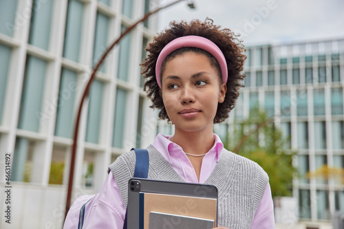 Horizontal shot of pensive millennial girl has curly hair holds notepads digital tablet carires backpack wears casual clothes poses against blurred city background duing daytime. University student photo