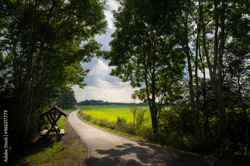 A narrow paved road next to agricultural fields near the city of Brandenburg. It's a sunny autumn day, but there are ominous storm clouds in the sky. There is a wooden bench on the left.