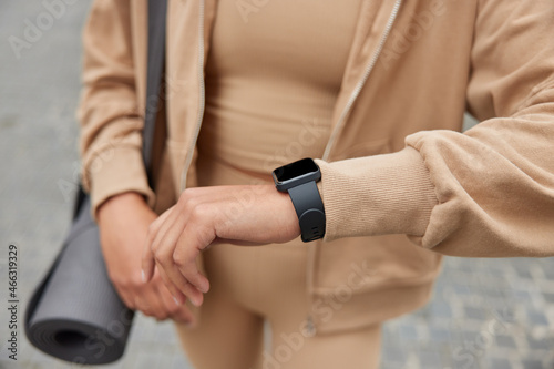 Cropped shot of faceless young sporty woman checks fitness results on modern smartwatch uses necessary application carries rolled up karemat dressed in sportswear poses against blurred background