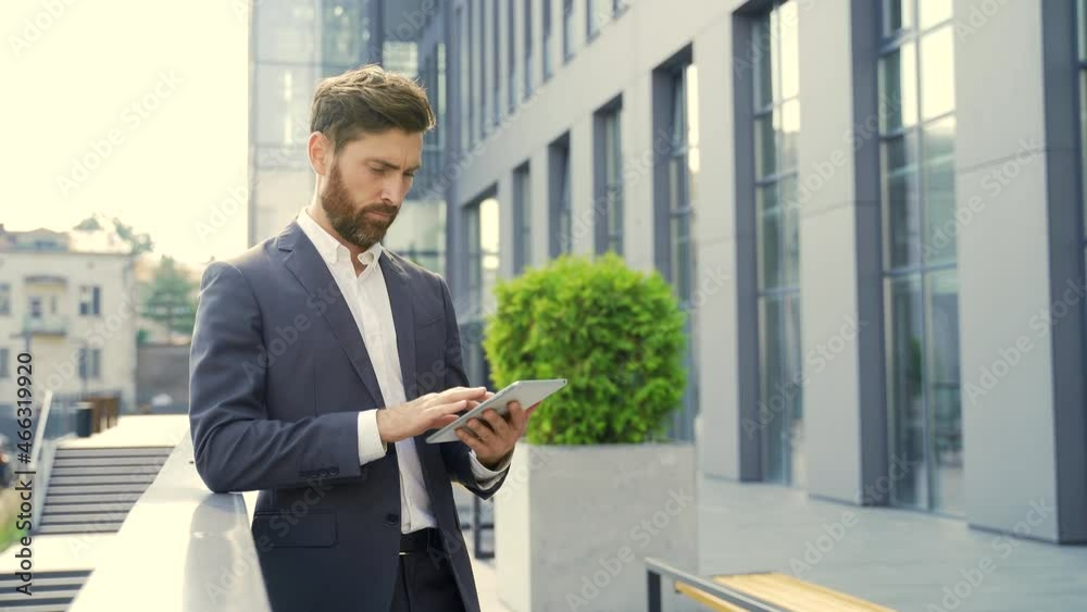 stylish bearded businessman in formal business suit standing working with tablet in hands on background modern office building outside. Man using smartphone or uses mobile phone outdoors city street stylish bearded businessman in formal business suit standing working with tablet in hands on background modern office building outside. Man using smartphone or uses mobile phone outdoors city street
