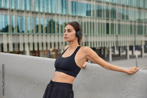 Active sporty woman dressed in sportswear does stretching exercises outdoors trains for having better body shape listens motivational music via headphones poses in urban setting. Sporty lifestyle