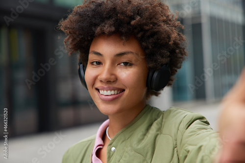 Photo of good looking cheerful woman with curly hair white teeth makes selfie during street walk listens music in headphones enjoys spare time favorite playlist. People and lifestyle concept