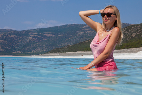 Young caucasian woman in the sunglasses and pink dress seating sexy posing in the Lake Salda, Turkey. Beautiful female happy relax walking on beach near sea with white sand and very clean blue water