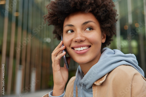 Positive woman with curly hair makes phone call keeps smartphone near ear concentrated away dressed in hoodie discusses something with friend during telephone talk stands outdoors uses cellular app