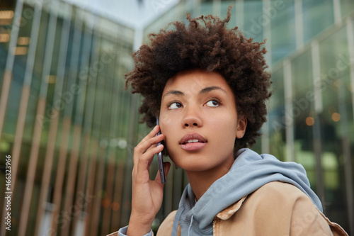 Serious curly haired woman listens received smartphone information during cell conversation dressed in hoodie looks thoughtfully away stands against blurred glass city building. Communication