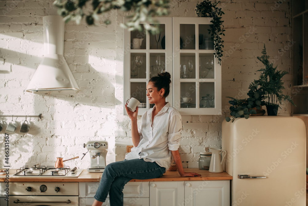 Smiling young brunette girl with long hair in a white shirt and jeans sits at home on the kitchen table with a mug in her hands Smiling young brunette girl with long hair in a white shirt and jeans sits at home on the kitchen table with a mug in her hands