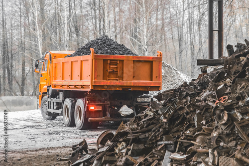 A dump truck in an industrial area or on a construction site in winter unloads coking coal from the body, next to a pile of metal waste and garbage