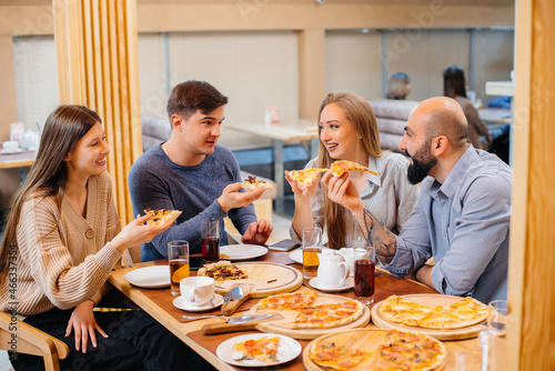 A group of young cheerful friends is sitting in a cafe talking and eating pizza. Lunch at the pizzeria.