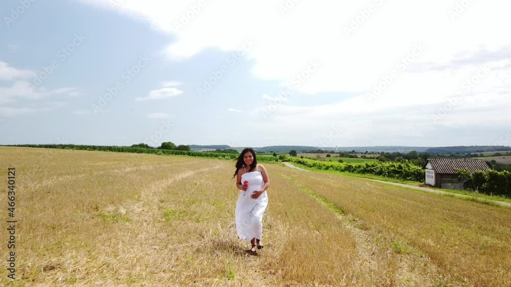 Young pregnant Woman in a White Summer Dress walking down a field holding her belly and flowers for a Maternity Photo Shoot Young pregnant Woman in a White Summer Dress walking down a field holding her belly and flowers for a Maternity Photo Shoot
