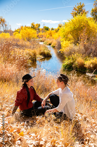 Lesbian couple smiling at each other with a river in a field during autumn surrounded by leaves.