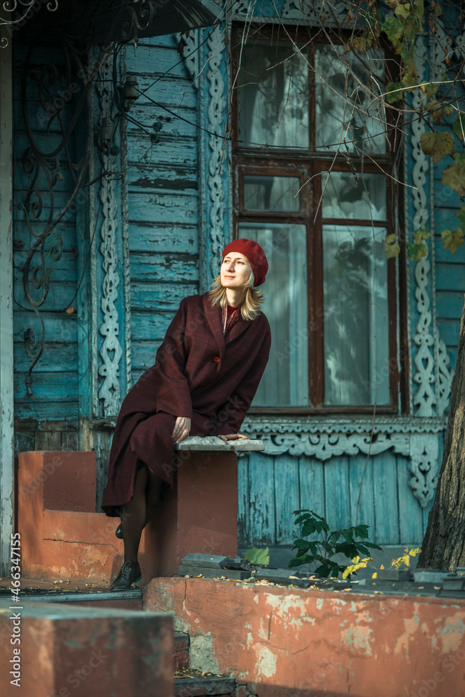 A woman in a burgundy coat sits on the porch of an old wooden house.