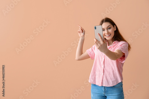 Young smiling woman taking selfie and pointing at something on beige background