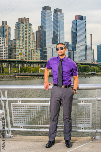 Dressing in a purple shirt, gray pants, a black tie, leather shoes, wearing a sunglasses, a young handsome businessman with a little beard and mustache is standing outside business district, relaxing. © Alexander Image