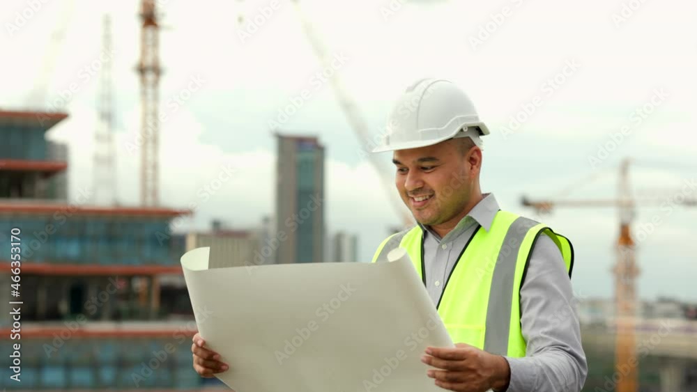 Asian engineer handsome man or architect looking blueprint with white safety helmet in city construction site . Standing with paperwork on rooftop building construction at capital. Asian engineer handsome man or architect looking blueprint with white safety helmet in city construction site . Standing with paperwork on rooftop building construction at capital.