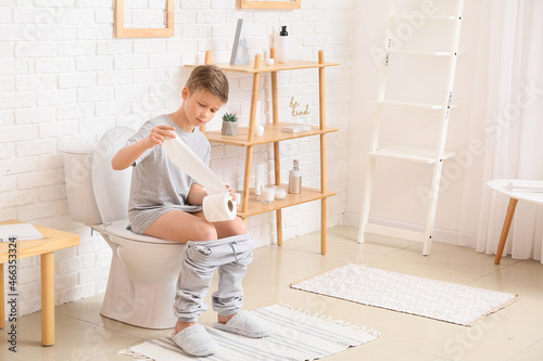 Little boy with paper sitting on toilet bowl in bathroom