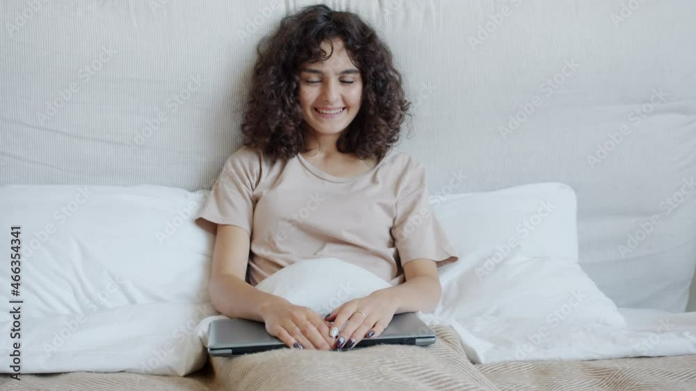 Slow motion portrait of happy young woman holding laptop smiling sitting in bed alone and looking at camera. Modern technology and bedroom concept. Slow motion portrait of happy young woman holding laptop smiling sitting in bed alone and looking at camera. Modern technology and bedroom concept.