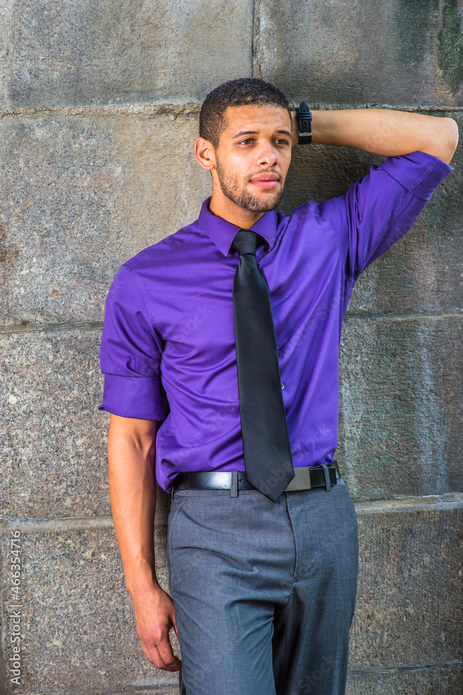 Obraz premium Dressing in a purple shirt, gray pants, a black tie, one hand putting in the back of his head, a young college student with a little beard and mustache is leaning against the wall, thinking outside..