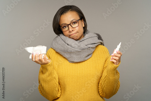 Unhealthy young woman with runny nose or sinusitis holding nasal drops and tissues. Unhappy black woman suffer from seasonal flu or allergy sneezing wear warm clothes and scarf. Influenza concept