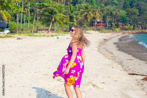 An adult blonde in a bright dress that sways in the wind, on the tropical seashore. Travel and tourism. © Natalia