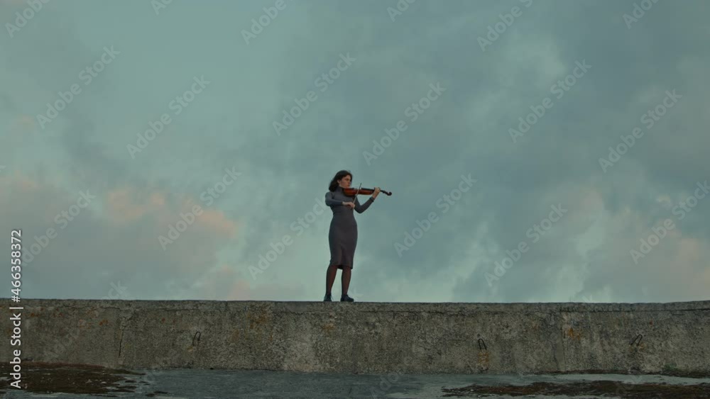 Lonely woman in grey dress is playing fiddle, standing outside, cloudy autumn sky is on the background, melancholic mood, Zoom in, Slow motion. Lonely woman in grey dress is playing fiddle, standing outside, cloudy autumn sky is on the background, melancholic mood, Zoom in, Slow motion.