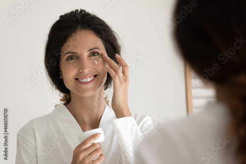 Latina woman in bathrobe stand in front of mirror hold jar apply anti aging cream on outer corners of eyes to reduce wrinkles, moisturize facial skin. Cosmetics caring of health beauty of skin concept