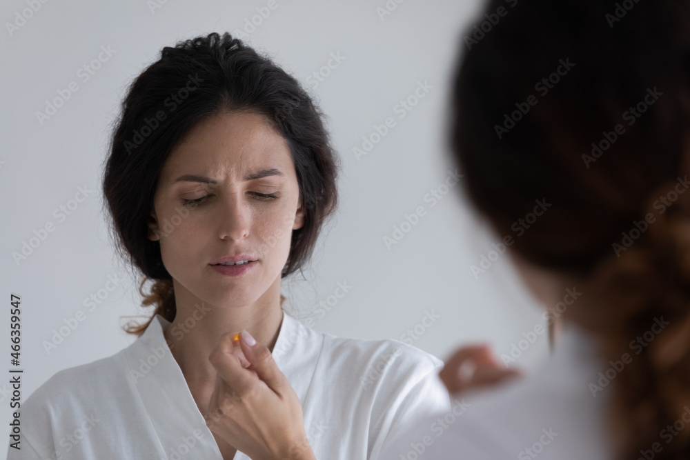 Young Hispanic woman in bathrobe holds pill in hand standing in bathroom reflecting in mirror. Frowning female feels unhealthy, takes capsule. Medical abortion decision, health-care, treatment concept Young Hispanic woman in bathrobe holds pill in hand standing in bathroom reflecting in mirror. Frowning female feels unhealthy, takes capsule. Medical abortion decision, health-care, treatment concept
