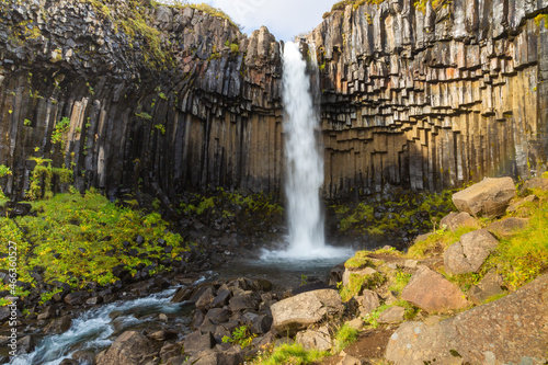 Color vertical photo of Svartifoss waterfall in Iceland. Blue sky with wispy clouds, basalt columns with orange highlights.
