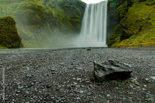 Fototapeta Naklejka Na Ścianę i Meble -  Closeup of Skogafoss waterfall in Iceland at sunrise.  Black and gray gravel in foreground. Green cliffs, no people.