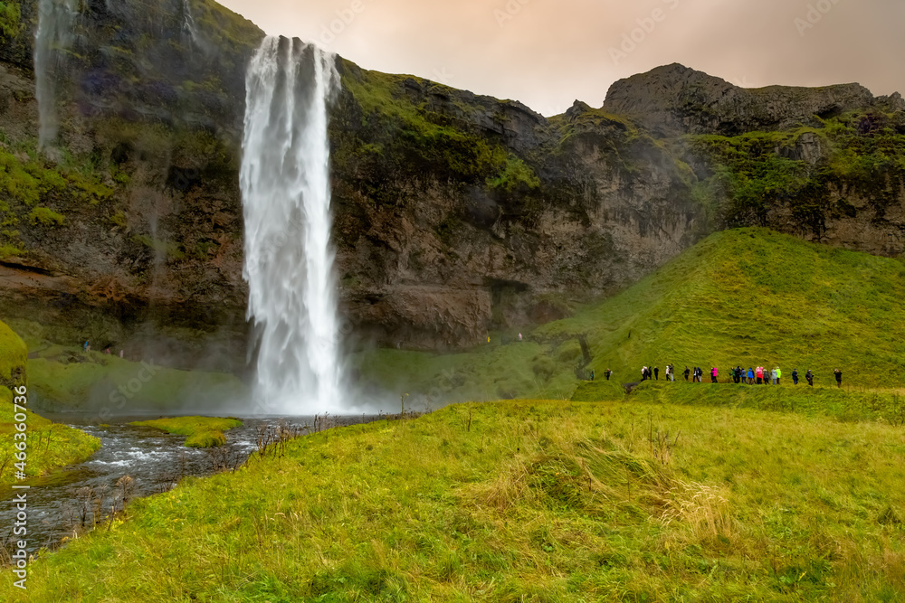 Seljalandsfoss waterfall at sunset in Iceland. Water is milky white set against verdant green cliffs with red and orange color in clouds. People visible on lower right of frame for scale. Seljalandsfoss waterfall at sunset in Iceland. Water is milky white set against verdant green cliffs with red and orange color in clouds. People visible on lower right of frame for scale.