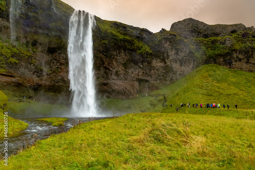 Seljalandsfoss waterfall at sunset in Iceland. Water is milky white set against verdant green cliffs with red and orange color in clouds. People visible on lower right of frame for scale.