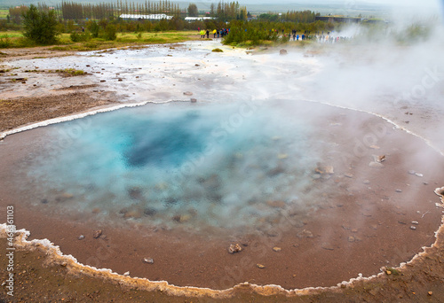 Strokkur geyser in Iceland. Geyser ring has a deep turquoise blue spot surrounded by red clay dirt with steam billowing out of the center of the ring.