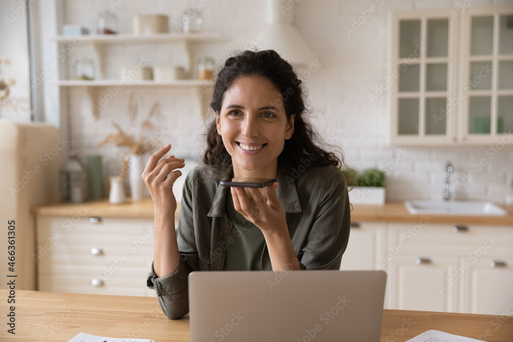 Smiling Latina woman sit at table with laptop holds smartphone talks on speakerphone, leave voicemail message chatting remotely enjoy personal or formal distant conversation. Modern ai tech concept - obrazy, fototapety, plakaty 