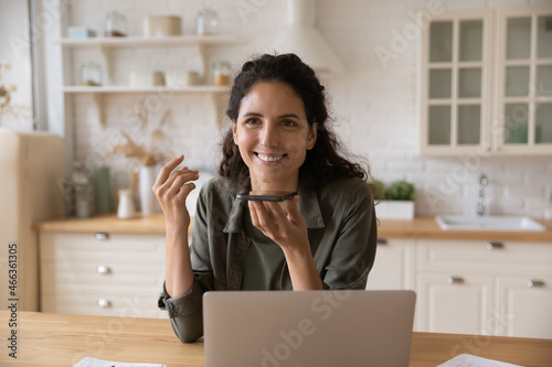 Smiling Latina woman sit at table with laptop holds smartphone talks on speakerphone, leave voicemail message chatting remotely enjoy personal or formal distant conversation. Modern ai tech concept