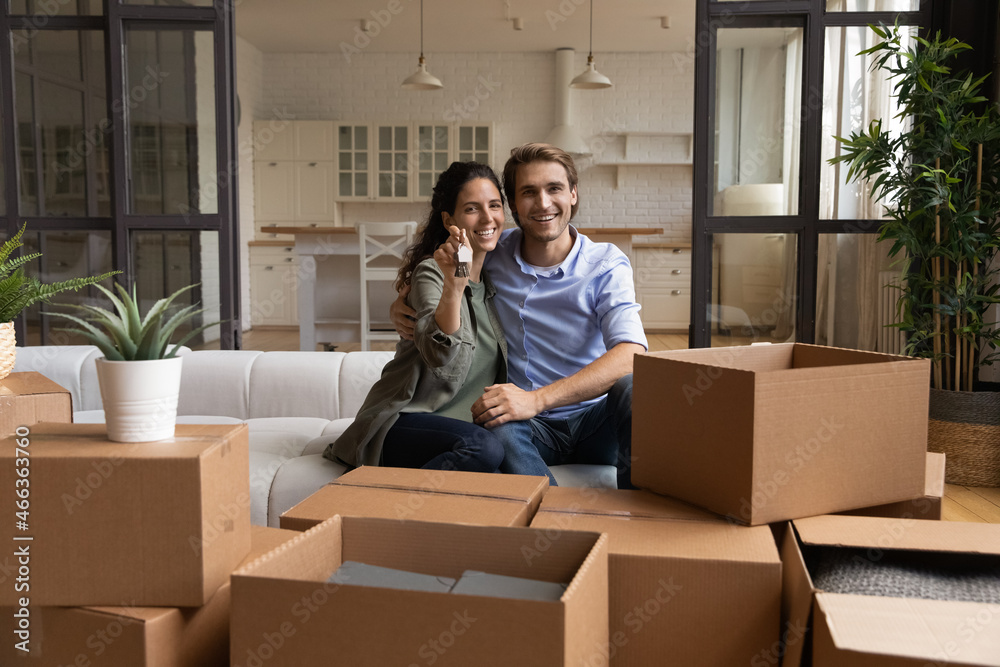 Happy couple enjoy relocation day, sit on sofa smile look at camera showing bunch of keys from new house, cardboard boxes with belongings nearby. Bank loan, tenant, homeowners family portrait concept Happy couple enjoy relocation day, sit on sofa smile look at camera showing bunch of keys from new house, cardboard boxes with belongings nearby. Bank loan, tenant, homeowners family portrait concept
