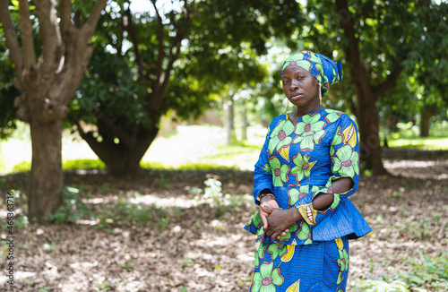 Worried lonely black woman looking sadly into the camera, standing among some trees in a West African village, wrestling hands in discomfort photo