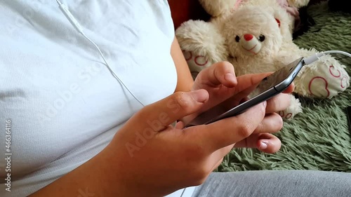 Happy girl in headphones listening music and using smartphone sliding screen, pressing with finger, surfing internet, social media, typing text. Relaxation in own room, via mobile phone in two hands